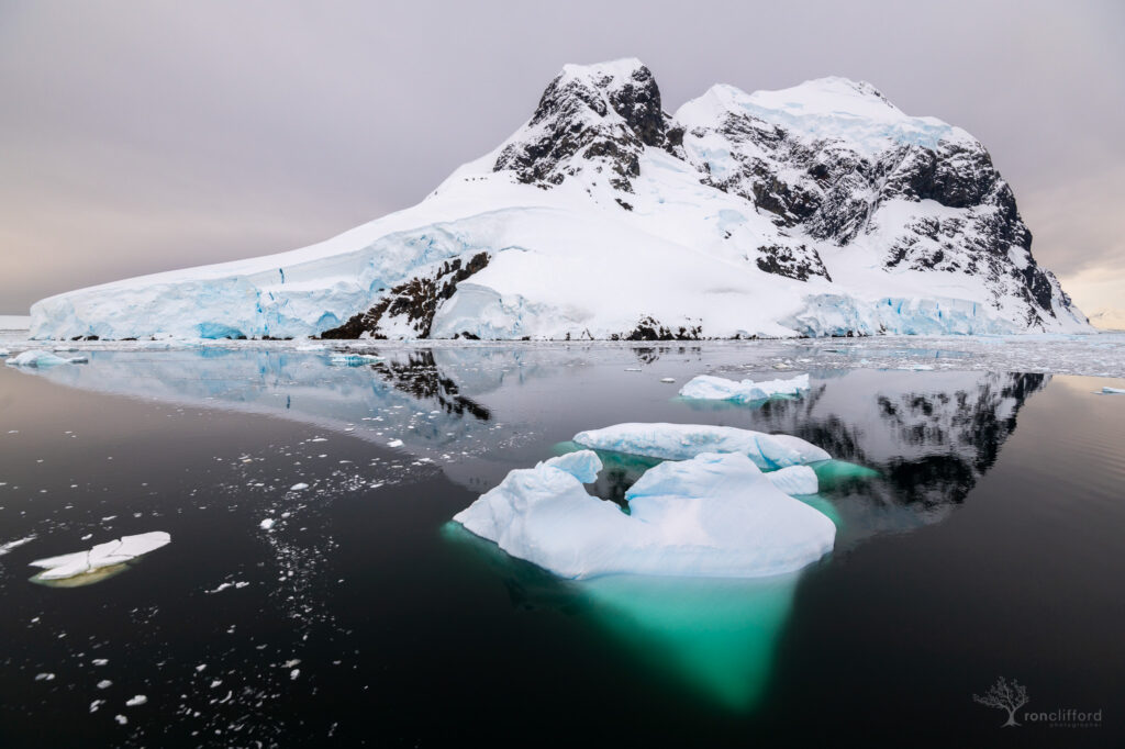 A glowing blue submerged iceberg in front of a mountain at the entrance of the lemaire channel